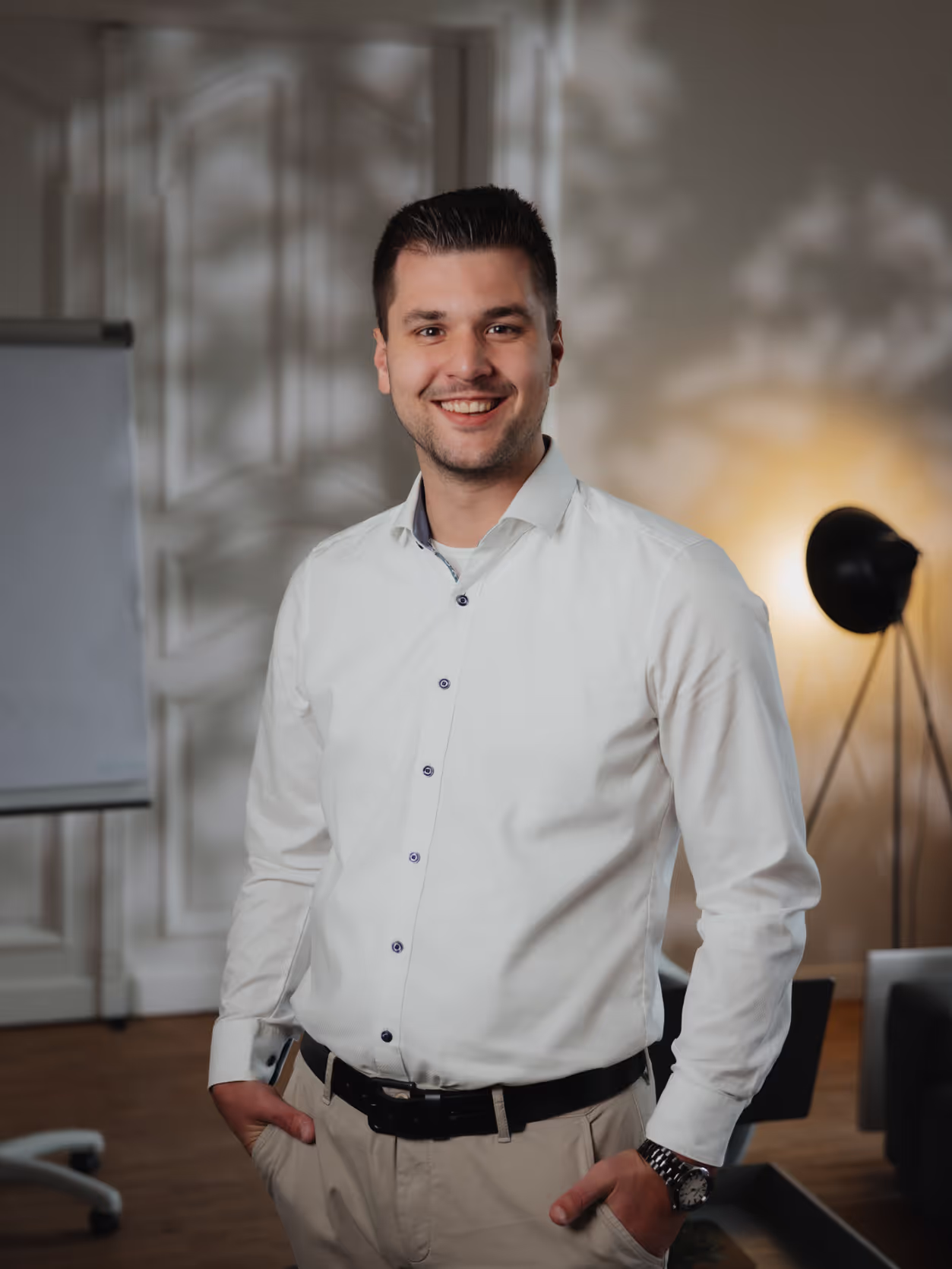 Junger Mann in weißem Hemd lächelt mit Händen in den Taschen in einem modern eingerichteten Büro.