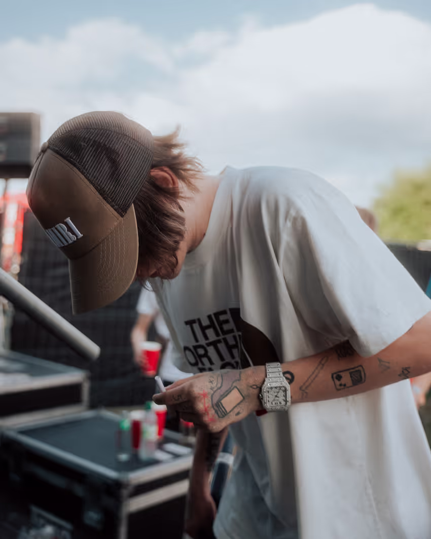 Ein junger Mann mit Baseballkappe und tätowierten Armen steht in einer lässigen Pose und blickt nach unten. Er trägt ein oversized T-Shirt mit einem Logo von “The North Face” und eine große Armbanduhr. Im Hintergrund sind verschwommene Gegenstände wie rote Becher und Equipment zu sehen, die auf eine Event- oder Konzertkulisse im Freien hindeuten. Die Szene wirkt entspannt und urban.