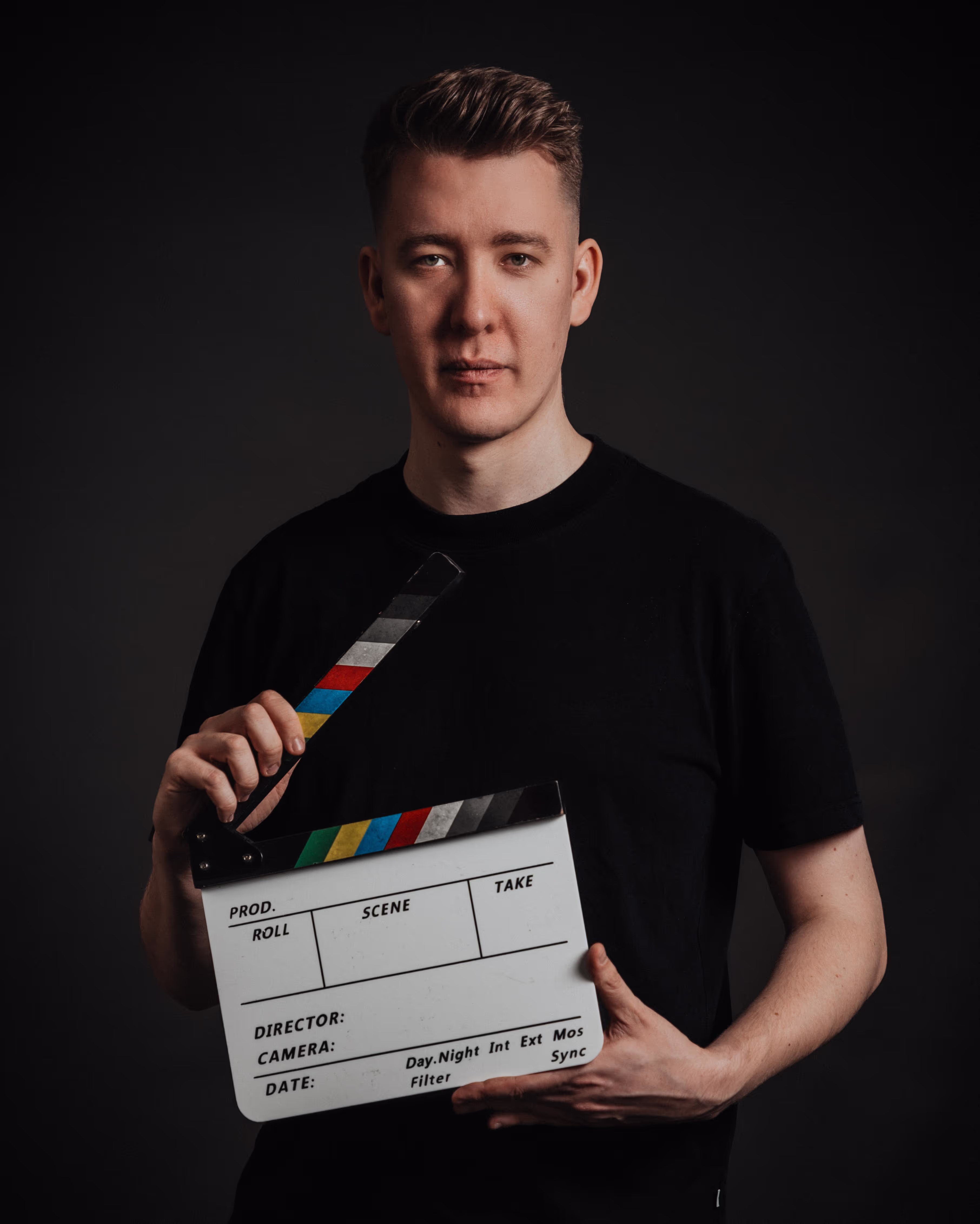 Ein Mann in schwarzer Kleidung hält eine Filmklappe vor einem dunklen Hintergrund. Er blickt direkt in die Kamera und vermittelt mit seiner Haltung Professionalität und Fokus. Die Filmklappe ist farbig markiert und symbolisiert seine Verbindung zur Filmproduktion. Die Szene ist minimalistisch beleuchtet, sodass der Fokus klar auf der Person und dem Objekt liegt.